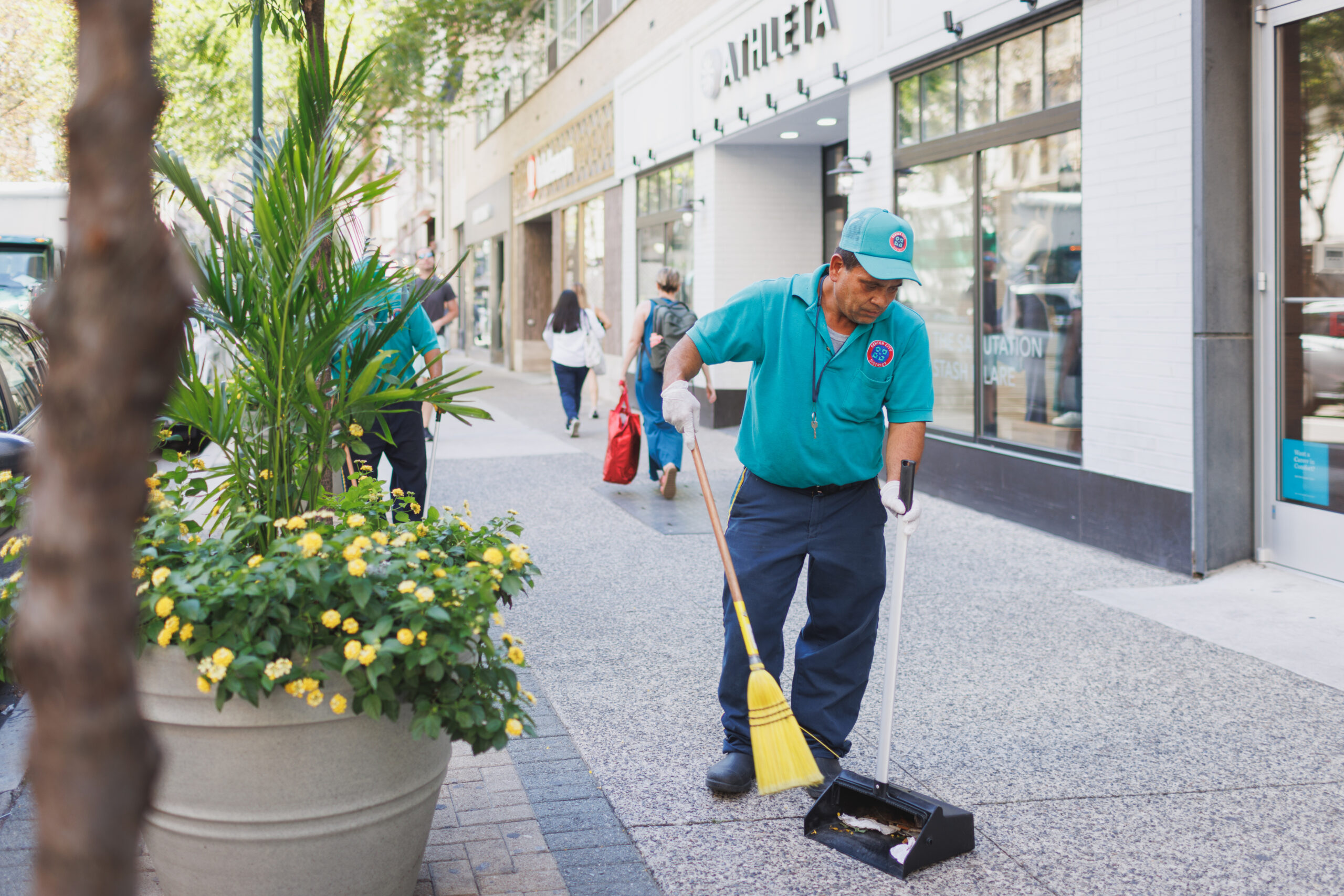 A CCD maintenance staff wearing a teal polo shirt and baseball cap sweeps the sidewalk with a pan and broom.