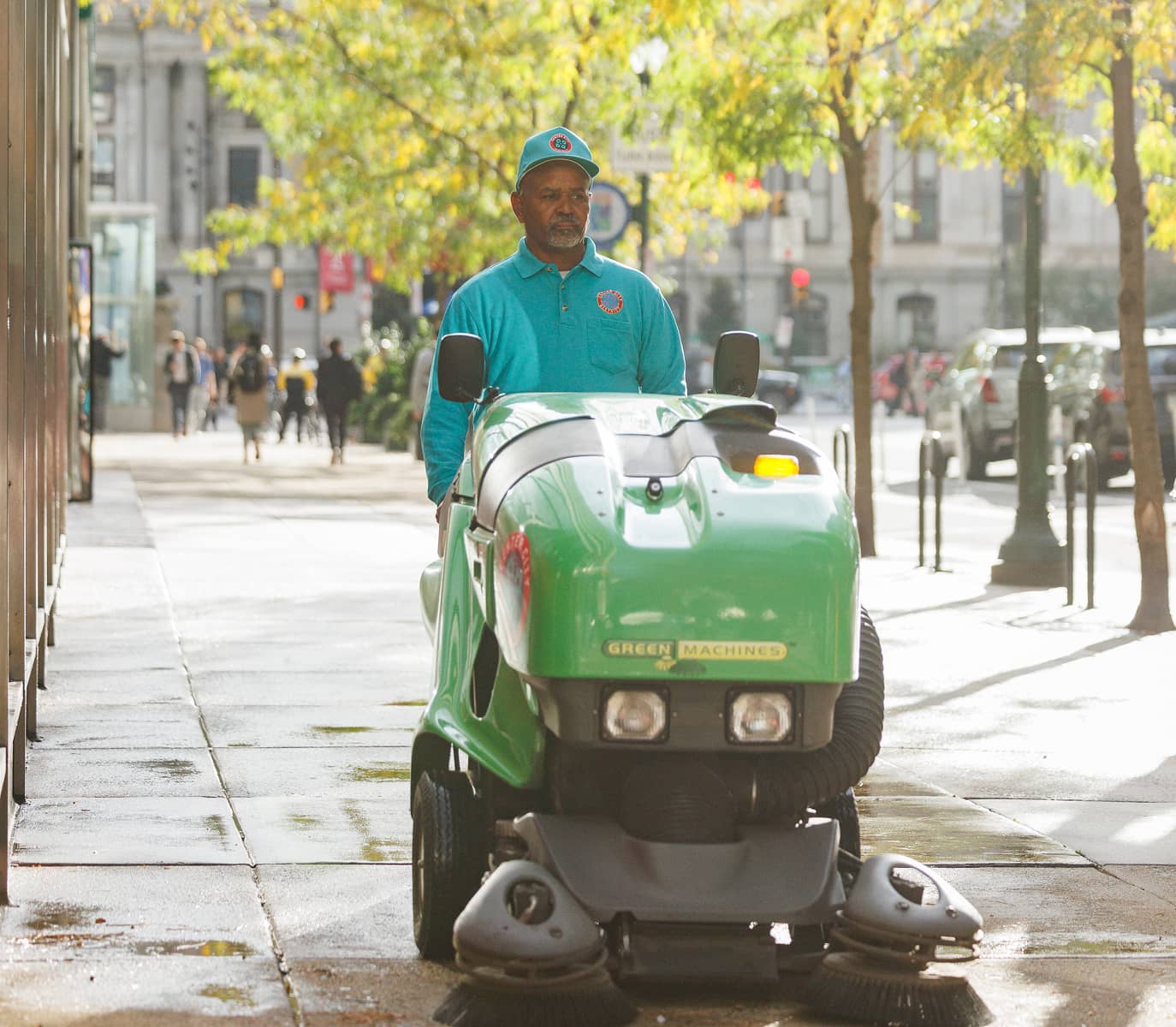 A CCD cleaner in a teal uniform operates a green street cleaning machine on a sunlit city sidewalk. Trees line the street, and pedestrians walk in the background.