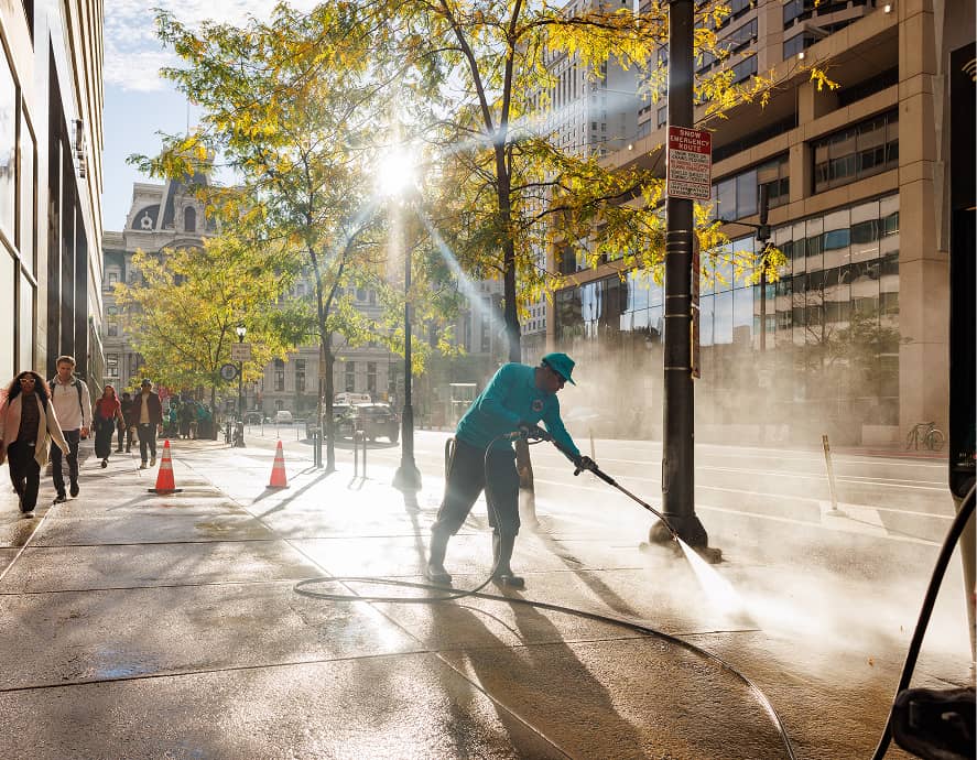 A CCD maintenance staff person in teal uses a hose to power wash a city sidewalk. Sunlight filters through trees, casting shadows. People walk by, framed by tall buildings.