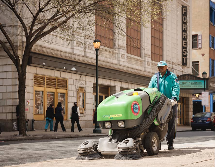 A CCD maintenance staff person operates a green machine, sweeping a city sidewalk. Nearby buildings and a theater marquee are visible, with pedestrians walking by.