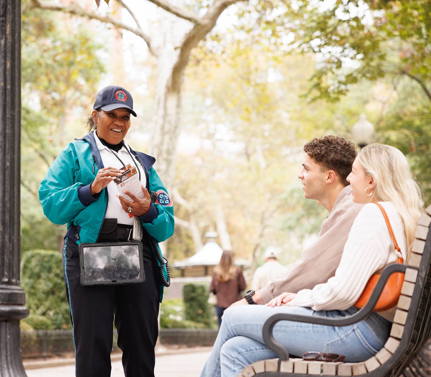 A CCD community service representative (CSR for short) safety ambassador wearing a uniform and carrying a 2-way radio, handing out brochures to a couple on a bench.