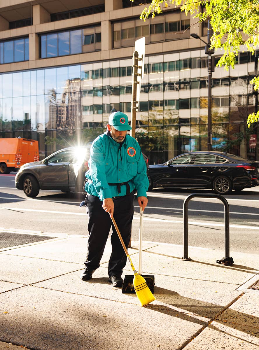 A CCD maintenance staff person in a teal uniform sweeps a sidewalk in bright sunlight. Behind him are parked cars, a modern glass building, and a tree with green leaves.