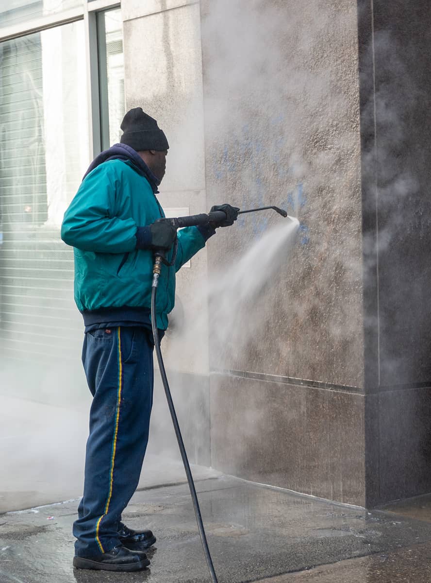 A CCD maintenance staff person in a teal jacket and hat power washes a graffitied wall on a city sidewalk. Dense mist and water spray create a focused, determined mood.