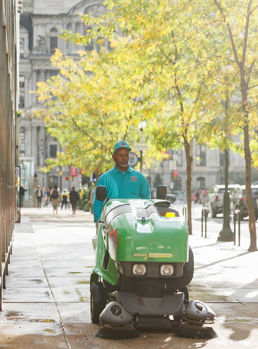 CCD maintenance staff wearing a teal polo shirt and baseball cap, pushing a green machine that cleans the sidewalk. Trees with yellow leaves and historic buildings line the background.