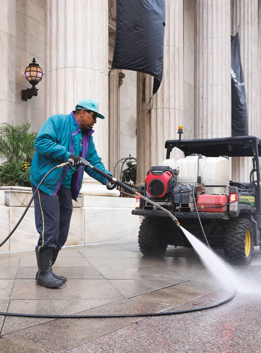 A CCD maintenance staff person wearing a teal jacket and cap power washes a sidewalk near a building with tall columns. The scene is industrious and focused.