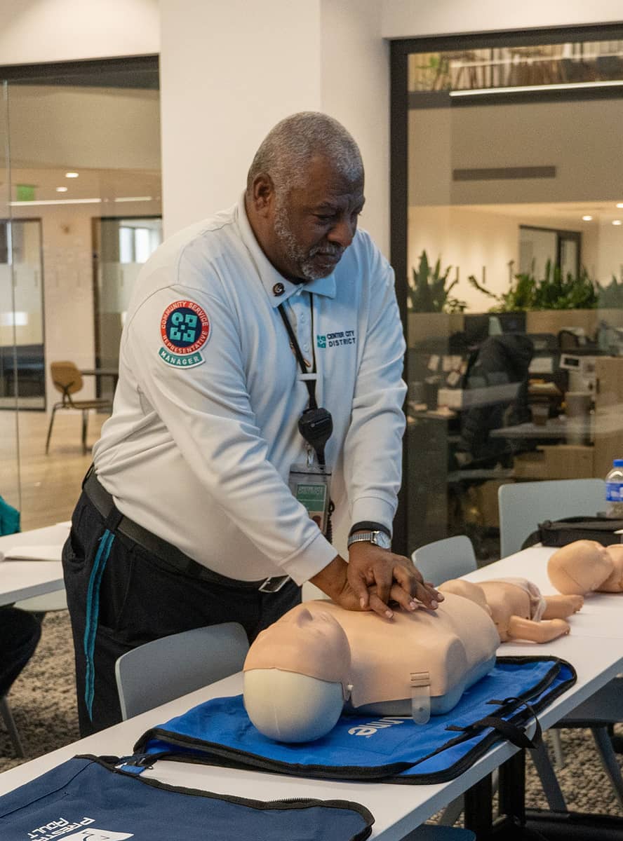 A CCD community service representative (CSR for short) supervisor wearing a uniform and carrying a 2-way radio, demonstrates CPR on a dummy during first aid training for a new class of CSR safety ambassador trainees.