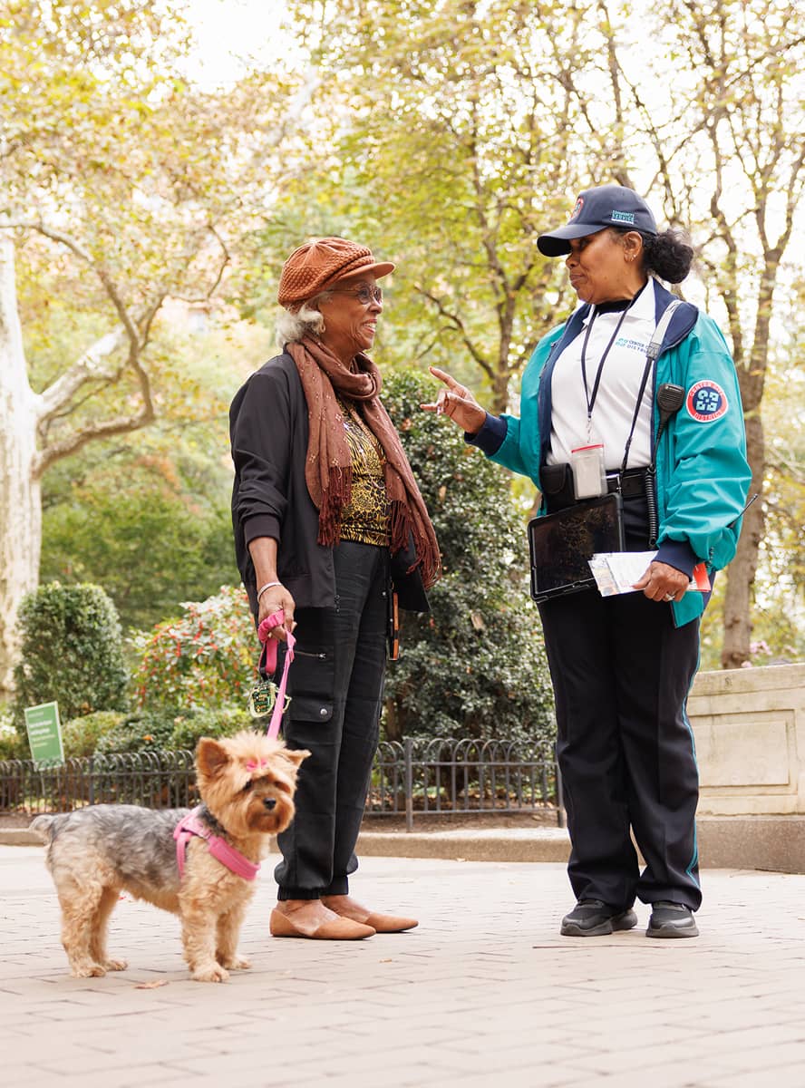 CCD community service representative (CSR for short) safety ambassador wearing a teal jacket, baseball cap and carrying 2-way radio and tablet, speaking to a woman who is walking her dog.