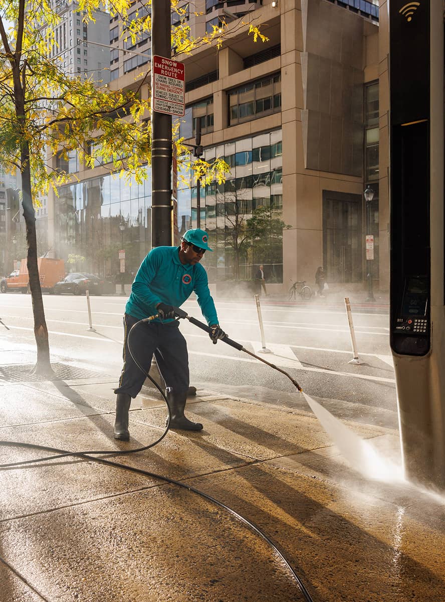 A CCD maintenance staff person in teal uses a hose to power wash a city sidewalk. Sunlight filters through trees, casting shadows. People walk by, framed by tall buildings.