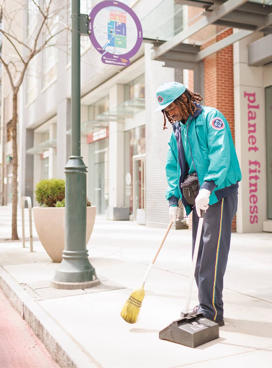 A CCD maintenance staff person in a teal uniform and hat sweeps a city sidewalk with a broom and dustpan. The street is clean and empty, suggesting a quiet, orderly setting.