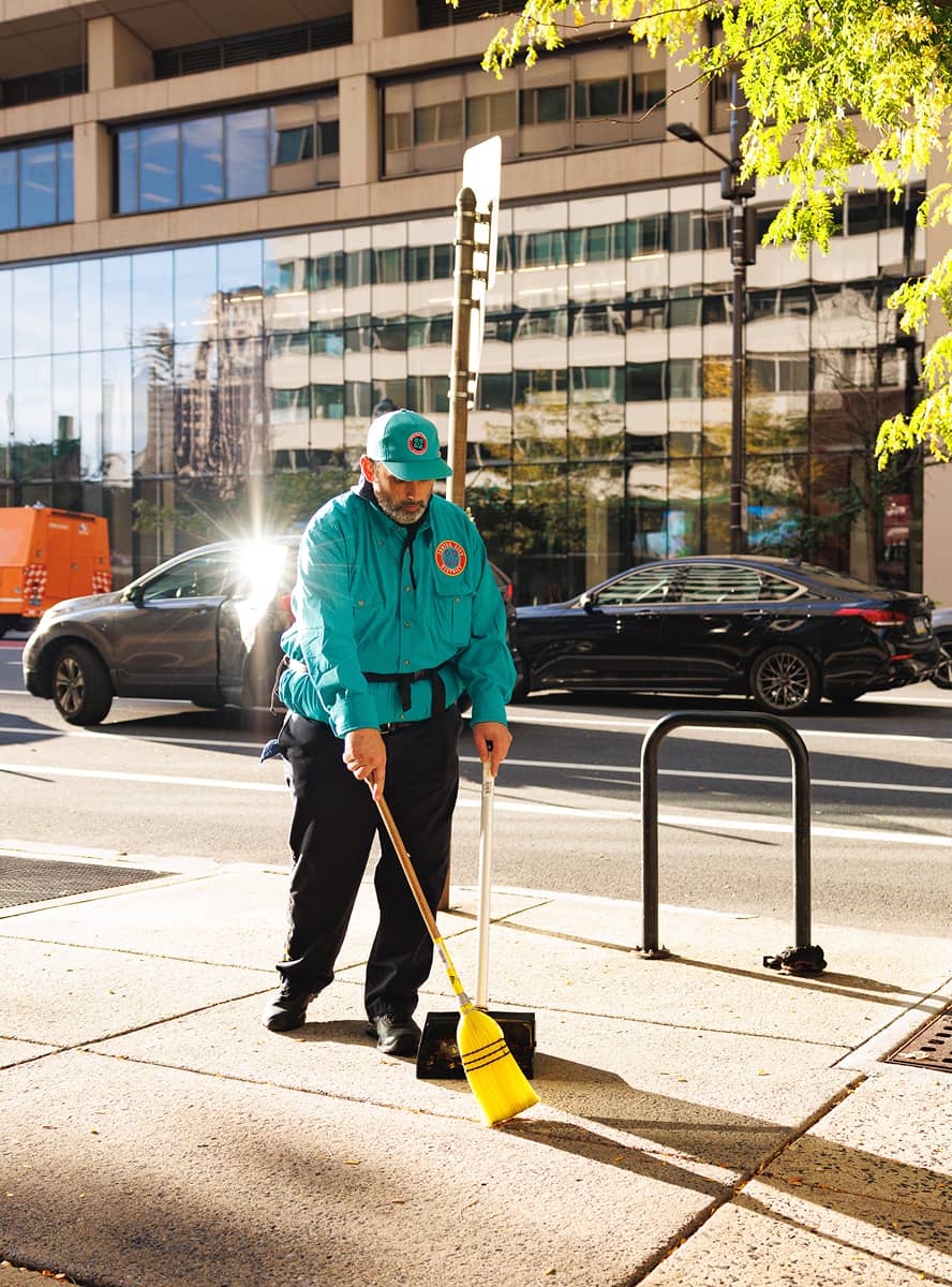 A CCD maintenance staff person in a teal uniform sweeps the sidewalk in bright sunlight. Behind is a busy street with parked cars and a glass-fronted building. The scene feels industrious and orderly.