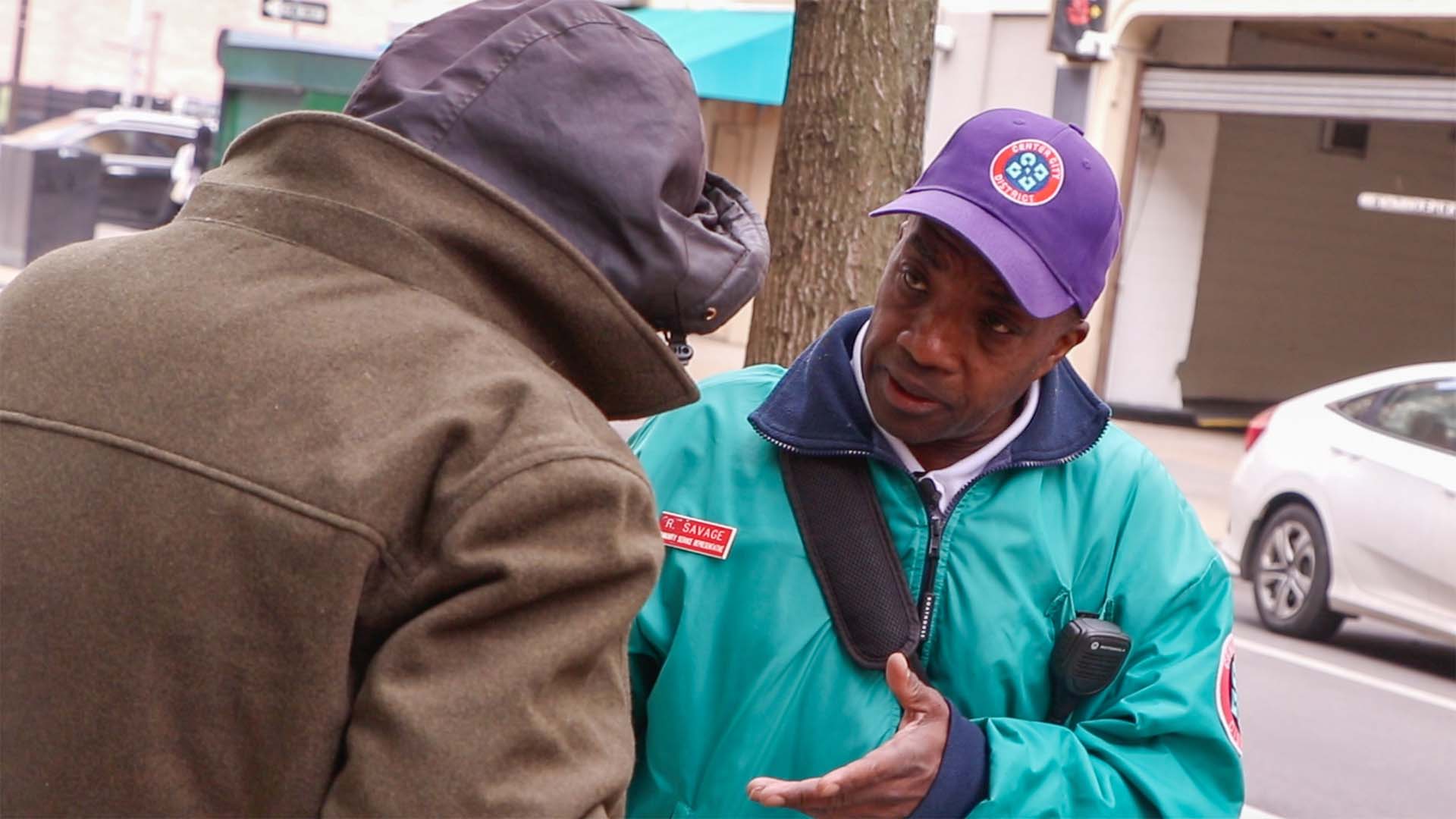 CCD community service representative (CSR for short) safety ambassador wearing a teal jacket, baseball cap and 2-way radio, helping an unhoused individual.