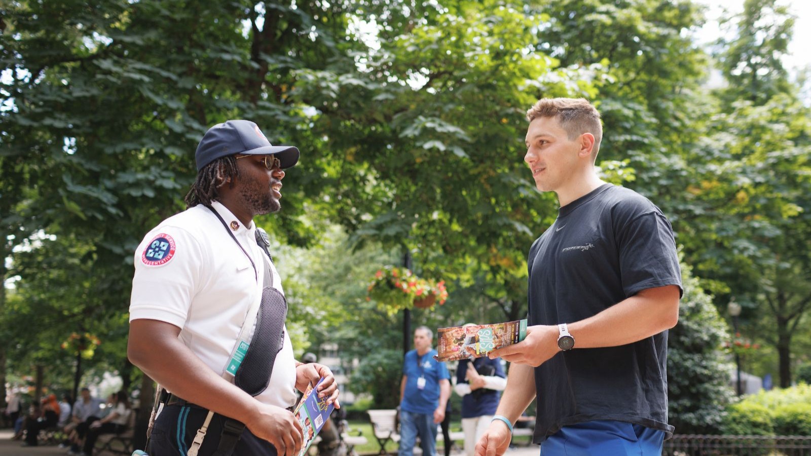 CCD community service representative (CSR for short) safety ambassador wearing a white uniform polo shirt, baseball cap and 2-way radio, talking to a person in front of a park.