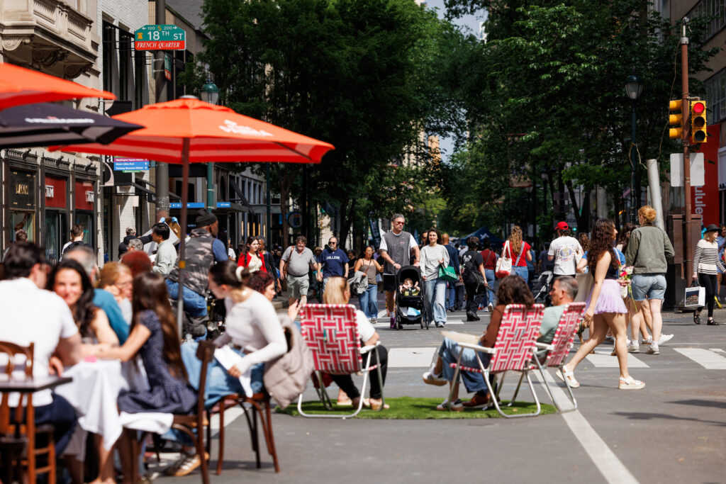 People sitting in the street at dining tables and on lawn chairs and people walking in the middle of the street .